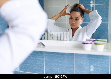 Die ziemlich dunkelhaarige Hispanic-Frau in weißem Bademantel bindet ihren Pferdeschwanz und schaut auf ihre Spiegelung im Badezimmerspiegel Stockfoto