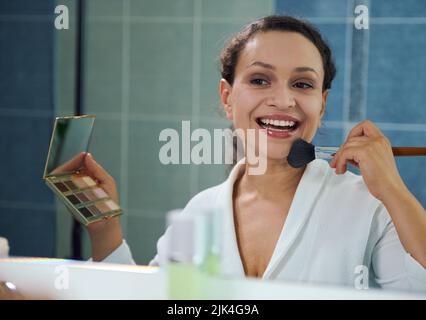 Spiegelung im Spiegel der entzückend lächelnden jungen Frau, hält Lidschatten Palette, trägt ein mattierendes Pulver auf ihr Gesicht Stockfoto