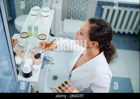 Draufsicht auf eine atemberaubende junge Frau, in einem weißen Bademantel hält bunte Lidschatten Palette und trägt Make-up auf ihr Gesicht Stockfoto