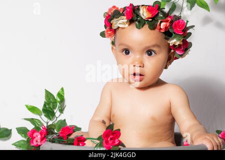 Nahaufnahme eines schönen braunen Latina-Mädchens, mit einem besorgten Gesicht und einer Krone aus Blumen, in einem Eimer, umgeben von Blumen. Mädchen, das wi sitzt Stockfoto