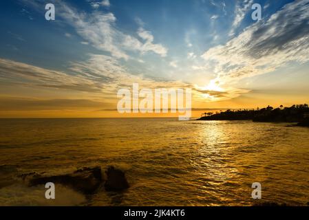 Laguna Beach Sonnenuntergang mit Wellen, die auf Felsen krachen und goldenen Reflexen auf dem Wasser. Stockfoto