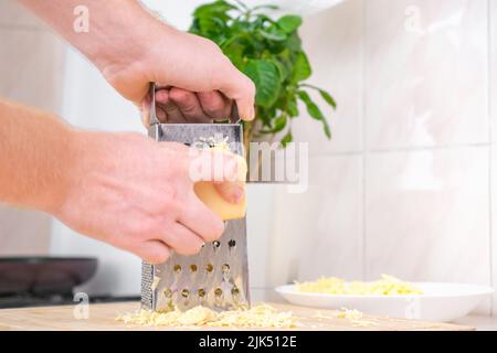 Ein Mann reibt Käse auf einer Metallreibe zum Einfrieren und zum weiteren Kochen von Pizza, Pasta. Stockfoto