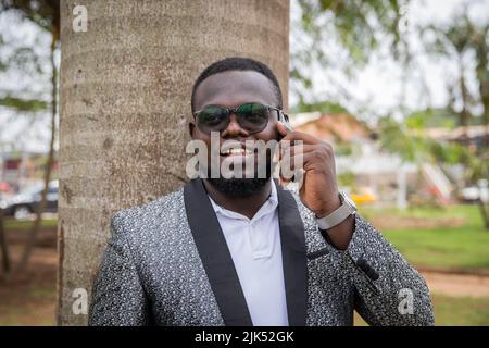 Ein junger lächelnder schwarzer Mann im Urlaub, der sich an einen Baum lehnt, ruft an. Stockfoto