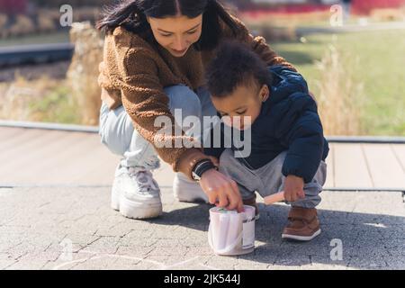 Eine schöne Mutter und ihr afrikanisches kleines Kind tragen stilvolle Winterkleidung und zeichnen auf dem Bürgersteig mit bunten Buntstiften - Park Hintergrund. Hochwertige Fotos Stockfoto