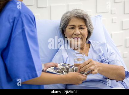 Ein älterer asiatischer weißhaariger Patient erhält von einer Krankenschwester Medikamente und ein Glas Wasser. Eine ältere Frau, die in einem Krankenhaus behandelt wird und auf dem Bett liegt Stockfoto