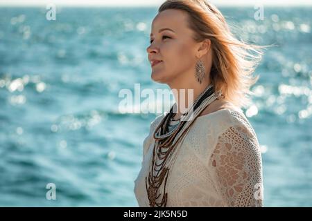 Frau in böhmischer Kleidung am Strand bei Sonnenuntergang. Boho-Stil für modischen Look im Resort. Frau mittleren Alters gut aussehende Frau in weißem Kleid und Boho-Stil Stockfoto
