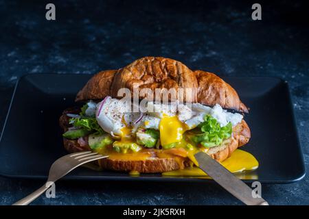 Croissant frisches Sandwich mit pochiertem Ei, Lachs und Avocado auf einem Teller auf schwarzem Hintergrund, Nahaufnahme Stockfoto