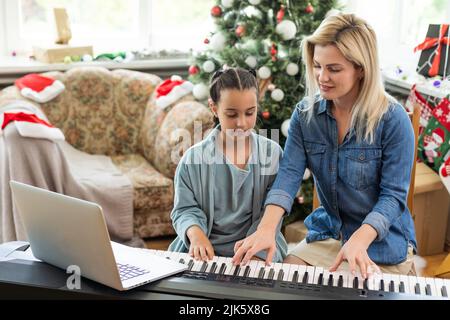Mutter lehrt ihre Tochter, am heiligabend in einem dekorierten Raum, Neujahrsstimmung, ein Neujahrslied auf dem Klavier zu spielen Stockfoto