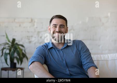 Glücklicher, gutaussehender Mann, der auf der Couch sitzt und die Kamera anschaut Stockfoto