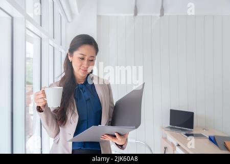 Geschäftsfrau trinkt eine Tasse Kaffee und steht an einem Fenster im Büro, Morgenarbeit im Büro Stockfoto