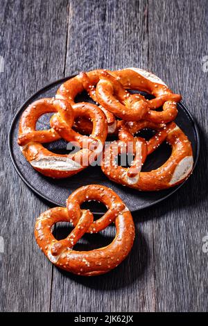 Weiche Brezeln in Form von Knoten gebacken und mit Salz auf schwarzem Teller auf dunklem Holztisch bestreut, vertikale Ansicht von oben, Nahaufnahme Stockfoto