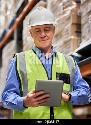 Ohne harte Arbeit kann es nicht gelingen. Ein reifer Mann, der ein digitales Tablet benutzt, während er in einem Lager arbeitet. Stockfoto