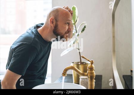 Junger erwachsener bärtiger Mann, der im Badezimmer im Spiegel sah und den Kopf berührte, sorgte sich um Haarausfall Stockfoto