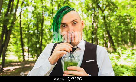 Junger Mann mit Dreadlocks trinkt erfrischende Limonade im Waldpark. Lustiger Hipster mit grüner Frisur trinkt Softdrink durch Stroh, abschreckend Stockfoto
