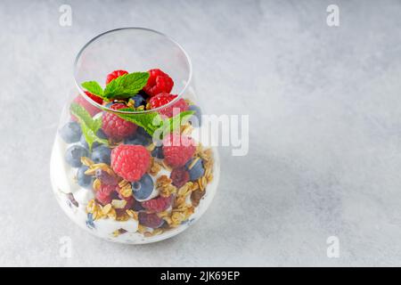 Müsli mit Joghurt und frischen Beeren im Glas serviert. Glas mit Puffdessert aus Naturjoghurt auf grauem Hintergrund. Gesundes Frühstückskonzept Stockfoto