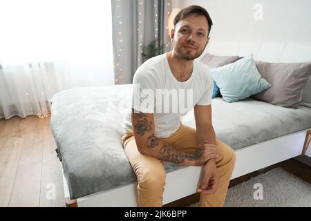 Lächelnder junger Mann sitzt auf dem Bett im Schlafzimmer Stockfoto
