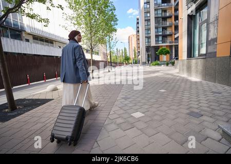 Eine Frau in einem Hijab geht mit einem Koffer auf Rädern die Straße hinunter Stockfoto