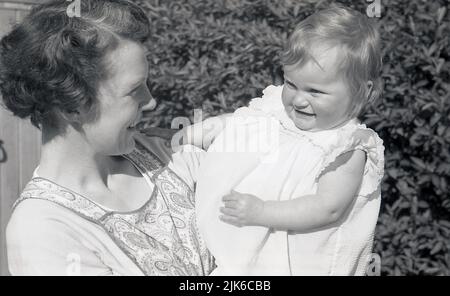 1960s, historisch, draußen im Vorgarten des Hauses, eine Mutter, die eine gemusterte Schürze der Zeit trug, ihre kleine Tochter in der Hand hielt und sie liebevoll ansah, England, Großbritannien. Stockfoto