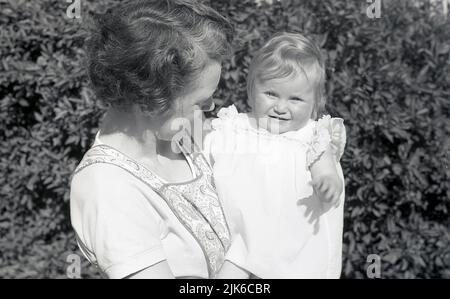 1960s, historisch, draußen in einem Garten, eine Mutter, die eine gemusterte Vorgeschichte der Zeit trägt, ihre kleine Tochter hält und sie liebevoll anschaut, England, Großbritannien. Stockfoto