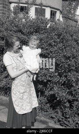 1960s, historicla, draußen, eine Mutter in einem Schürzenkleid, die ihr Kleinkind in der Hand hält, England, Großbritannien. Stockfoto