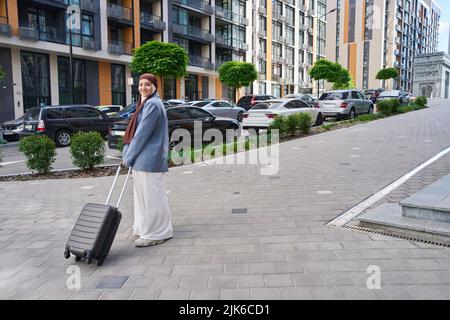 Frau steht auf einer verlassenen Straße und hält einen Koffer auf Rädern Stockfoto