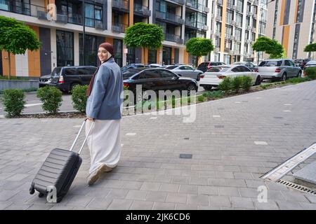 Frau steht in einer halben Umdrehung und hält einen Koffer auf Rädern Stockfoto