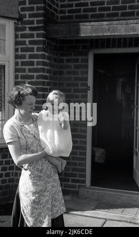 1960s, historisch, vor dem Eingang zu einem Haus, eine Mutter, die einen gemusterten Vorbau der Zeit trug und ihre kleine Tochter, England, Großbritannien, in der Hand hielt. Stockfoto