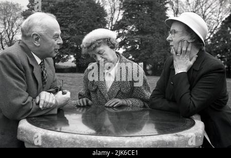 1960s, historisch, draußen in einem Park, drei gut gekleidete ältere Menschen, die auf dem Messinggesicht einer steinernen Sonnenuhr gelehnt sind, plaudern, England, Großbritannien. Stockfoto