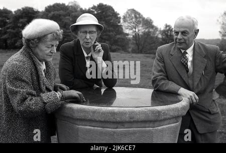 1960s, historisch, draußen in einem Park, drei gut gekleidete ältere Menschen, die auf dem Messinggesicht einer steinernen Sonnenuhr gelehnt sind, plaudern, England, Großbritannien. Stockfoto