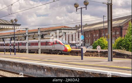 Eine moderne Lokomotive steht neben einem historischen Vordach mit einem Backsteingebäude dahinter. Schienen und eine Plattform stehen im Vordergrund und ein wolkiger Himmel ist oben. Stockfoto