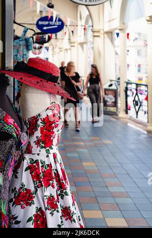 Rote Rosen Kleid mit rotem Hut und schwarzem Band in Mode-Shop in der High Street auf einem Manequin angezeigt. Die Einkäufer im Hintergrund laufen durch die Arkade. Stockfoto