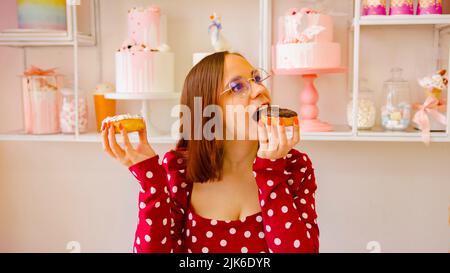Eine schöne Frau in einem roten Kleid und einer Brille hält einen weißen Donut in der einen Hand und beißt einen Schokoladendonut, während sie ihn mit der anderen Hand im c hält Stockfoto