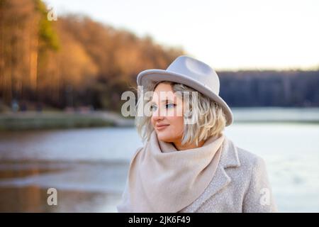 Porträt einer blonden Frau in einem Hut mit hellen Augen Make-up im Herbst in den Strahlen der untergehenden Sonne vor dem Hintergrund der Natur - Wasser und Fores Stockfoto