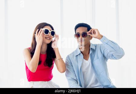 Glückliches asiatisches Paar im Sommer, junger Mann und Frau mit Sonnenbrille und Lächeln, sitzen im hellen Hotelzimmer in der Nähe des weißen Vorhangs und des Glases Stockfoto