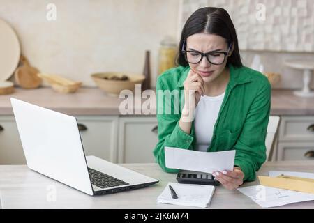 Schöne Frau in einem grünen Hemd in Verzweiflung, erhielt einen Brief mit schlechten Nachrichten, Brünette liest eine Nachricht mit Traurigkeit in der Küche zu Hause Stockfoto