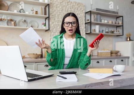 Junge schöne Frau in Brille trägt grünes Hemd Blick auf Kamera enttäuscht und wütend auf Hausaufgaben Papierarbeit, hält rotes Telefon, Brünette Berechnung der Haushaltsfinanzen in der Küche Stockfoto