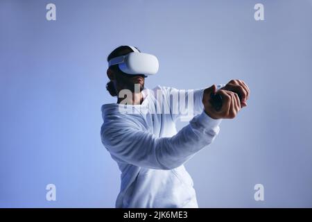 Mann spielt ein Cricket-Spiel in der virtuellen Realität. Sportlicher junger Mann, der mit Gaming-Controllern einen virtuellen Ball zugeschlagen hat. Aktiver junger Mann, der Immersi erkundet Stockfoto