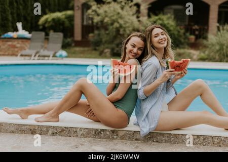 Zwei niedliche junge Frauen sitzen am Pool und essen Wassermellon im Hinterhof des Hauses Stockfoto
