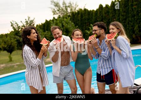 Gruppe junger Menschen, die am Pool stehen und im Hinterhof des Hauses Wassermellon essen Stockfoto
