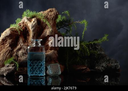 Blauer Gin auf einem schwarzen reflektierenden Hintergrund. Wacholderzweige, Stein und altes Treibholz stehen im Hintergrund. Stockfoto