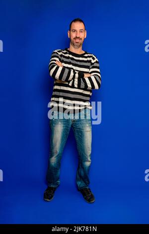 Lächelnder bärtiger spanischer Mann posiert mit gekreuzten Armen auf einem vertikalen Foto in voller Länge, isoliert auf blauem Studiohintergrund. Stockfoto