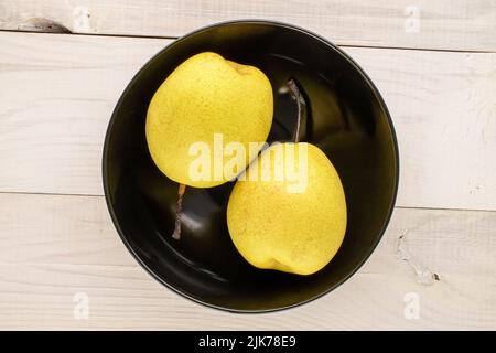 Zwei süße, leuchtend gelbe Birnen in einer schwarzen Keramikplatte auf einem Holztisch, Nahaufnahme, Draufsicht. Stockfoto