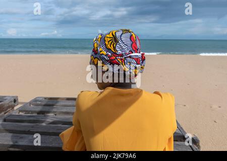 Ghanaische Frau am Strand mit afrikanischem, farbenfrohen Kopfschmuck und sitzt auf einer Holzbank im Keta-Dorf Stockfoto