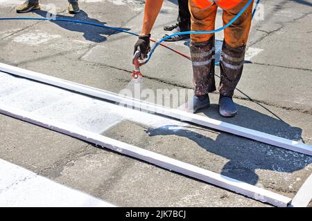 Ein Straßenarbeiter malt eine weiße Straßenmarkierung eines Fußgängerübergangs mit einer Holzrahmenschablone und einer Airbrush. Stockfoto