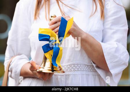 Beschnittenes Foto von weiblichen Händen, die die Schulglocke mit einer blau-gelben Schleife halten - ein Symbol für den Beginn des Schuljahres oder des Schulabschlusses in der Ukraine. Konz Stockfoto