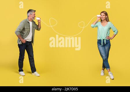 Kaukasischer Mann mittleren Alters, der seiner Frau durch Zinn etwas erzählt, kann mit gemalter Schnur, gelbem Hintergrund telefonieren Stockfoto