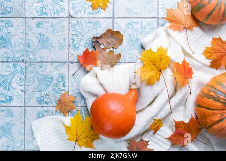 Kuschelige Herbst-Flachlage mit Kürbissen, gestricktem Wollpullover und Herbstlaub-Kerze auf Fliesenhintergrund. Einrichtung im Herbst. Gemütliche Herbststimmung. Erntedankfest. Halloween. Stockfoto