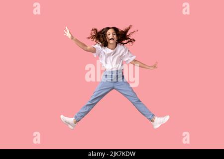 In voller Länge Porträt von aufgeregt liebenswert kleines Mädchen trägt weißes T-Shirt Springen in der Luft und spreizte die Arme, lächelnd glücklich, Spaß zu haben. Innenaufnahme des Studios isoliert auf rosa Hintergrund. Stockfoto