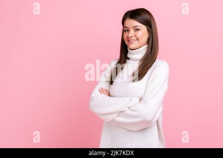 Positive selbstbewusste Frau mit dunklem Haar, die mit gekreuzten Armen steht, lächelnd auf die Kamera blickt und einen weißen Pullover im lässigen Stil trägt. Innenaufnahme des Studios isoliert auf rosa Hintergrund. Stockfoto