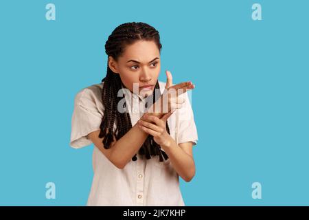 Portrait einer Frau mit schwarzen Dreadlocks, die mit einem Fingergewehr zeigen, mit Pistolenhandbewegung zielen und drohen, in einem weißen Hemd zu schießen. Innenaufnahme des Studios isoliert auf blauem Hintergrund. Stockfoto